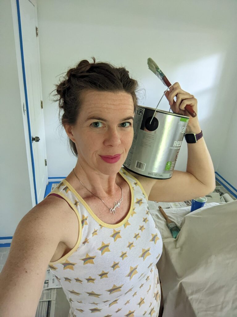 selfie of a person holding a can of paint and a brush with a bare room behind her