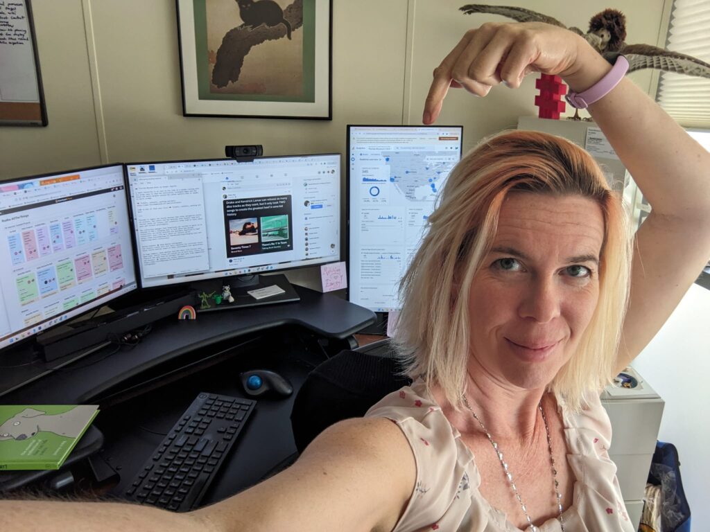 a person taking a selfie at a desk and pointing at a third monitor