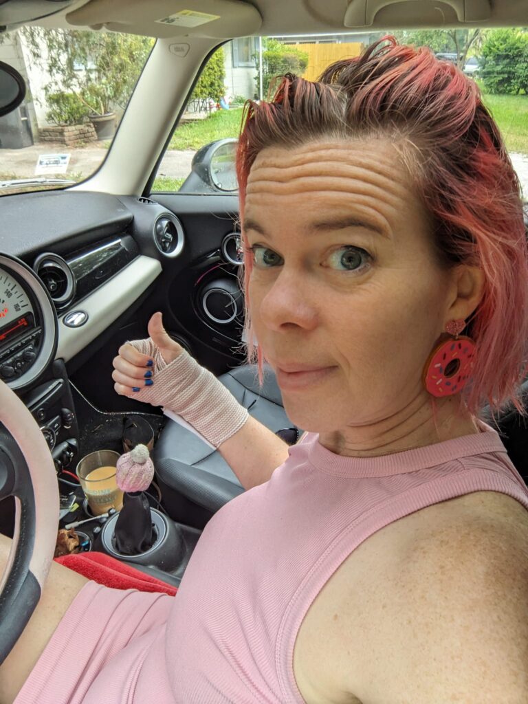 selfie of a person wearing a wrist bandage in a car making a thumbs up gesture over the gear shift