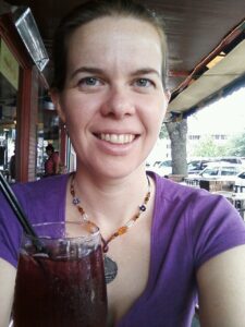 a woman wearing a purple shirt and a beaded necklace is smiling over a glass of dark beverage of some kind