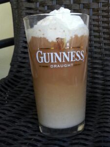 a Guinness pint glass seems to be full of coffee and cream and topped with whipped cream sitting on a dark woven surface