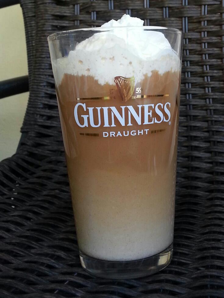 a Guinness pint glass seems to be full of coffee and cream and topped with whipped cream sitting on a dark woven surface