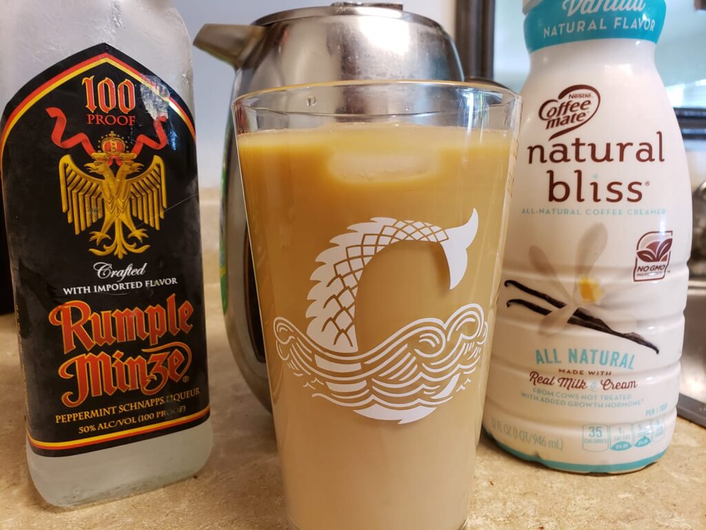 a pint glass with pale bwown liquid might be coffee because it is sitting in front of a coffee pot, a bottle of natural bliss creamer and a bottle of Rumple Minze liquor