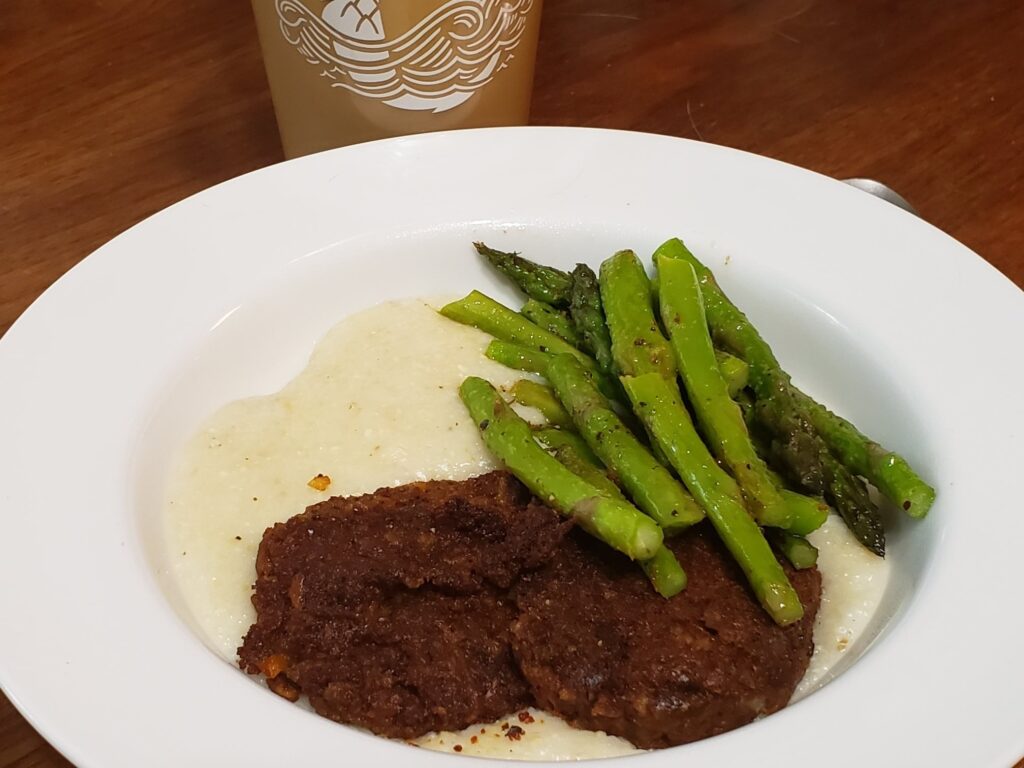 a white bowl has grits and asparagus and some kind of brown patties