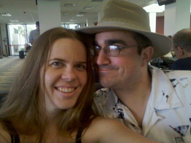 selfie of two people, one wearing a wide brimmed hat, in a public space like an airport or lobby