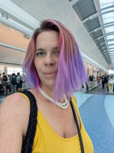 selfie of a person with purple hair standing in what looks like a busy public corridor in a mall or airport