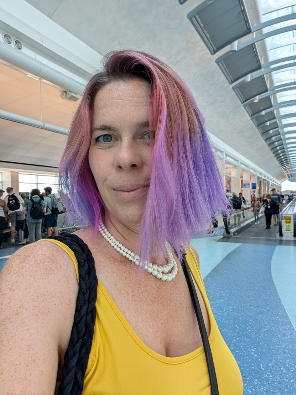 selfie of a person with purple hair standing in what looks like a busy public corridor in a mall or airport