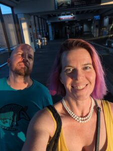 selfie of two people walking down a corridor in a public space with strong angled light on them as if the setting sun