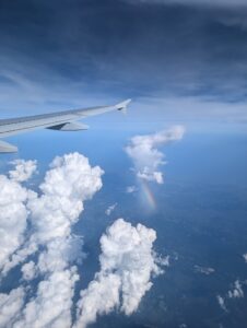 a view out a airplane window with part of the wing above columns of clouds and a faint rainbow
