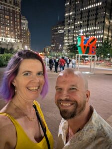 selfie of two people with a night time city scene behind them with tall buildings and a park with an iconic stylized statue of the stacked letters for LOVE
