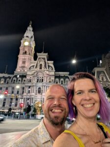 two people are taking a selfie with a very large elaborate building with a tall tower in the center against a night sky