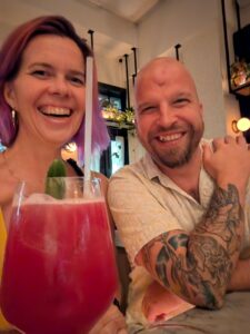 a selfie of two people in a bar with a pink cocktail in the foreground