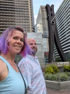 selfie of two people in an urban setting with a huge sculpture of a clothes pin in the background
