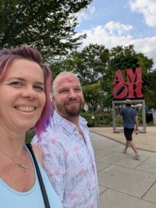 selfie of two people smiling in front of a metal statue of the stacked letters for AMOR