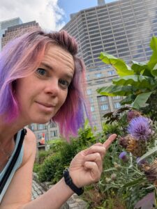 selfie of a person in an urban park crouching to point at a thistle flower that matches her purple hair