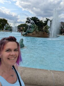 a person is smiling in front of a large water fountain with copper figures reclining in the middle