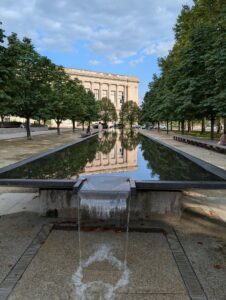 a long rectangular reflecting pool with a water fall at the near end is reflecting the rows of trees on either side and the squat square building in the distance