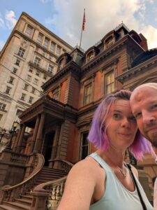 selfie of two people in front of an elaborate red and dark gray building with a wine curved stairway and another taller building towering behind it