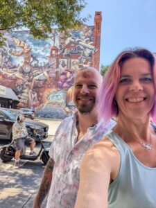 selfie of two people in dappled light with a building in the distance with a mural of a lot of chaotic symbolism interwoven on it