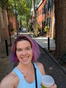 a person is smiling with a narrow street behind her lined with two story brick houses