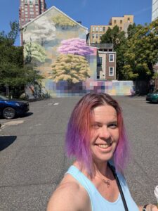 selfie of a person in bright sunlight in a parking lot with a building in the back covered in a mural of trees covered in flower blossoms