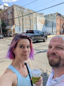 a selfie of two people with a city street behind them and beyond that is a soft colored mural of tall trees and snow on a building