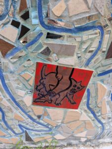 a wall of tiled mosaic has a central tile painted with a cat rubbing against someone's leg