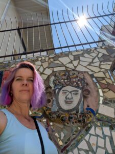 a person stans in front of a wall covered in tiled art and a painted face with sun shining from above through a metal fence railing