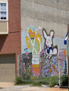 the side of a building behind a fence has a graffiti mural of a rabbit in a suit and a turtle in a jet pack