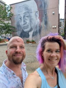 selfie of two people with a mural of a vintage photo of a man in the background on a building