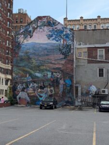 a parking lot buts up to a building with a mural on it of a view out over a wide valley and people in the foreground sitting on a balcony
