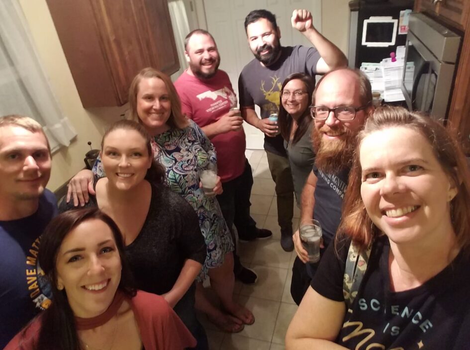 a group nine people are standing in two rows in a kitchen and smiling up at the camera