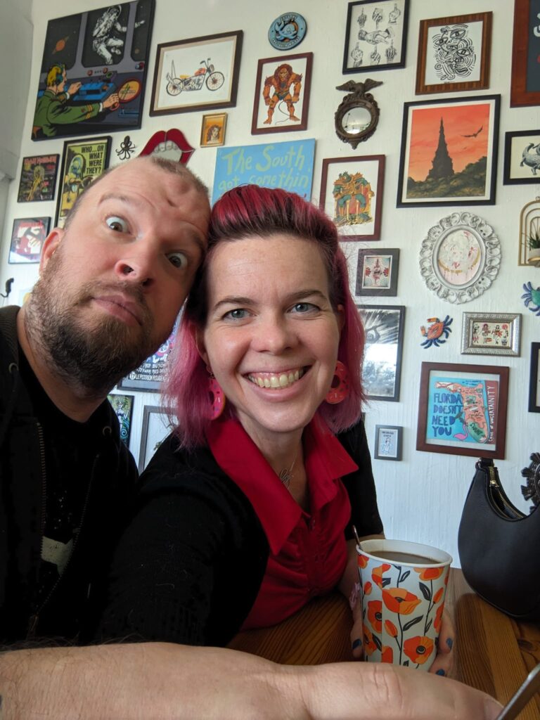 two people leaning in close to take a smiling selfie in front of a wall of many small prints on a wild variety of frames