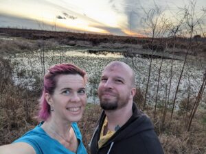 two people taking a selfie under a striking sunset being reflected in the wetland just behind them
