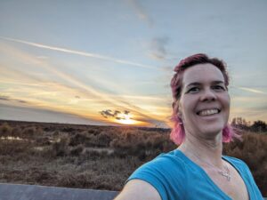 selfie of a smiling person with pink hair against a dramatic sunset over an open prairie