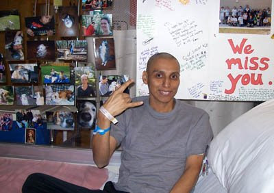 a bald person with hospital bracelets is sitting in a bed making a gesture with hand drawn posters and photos behind them