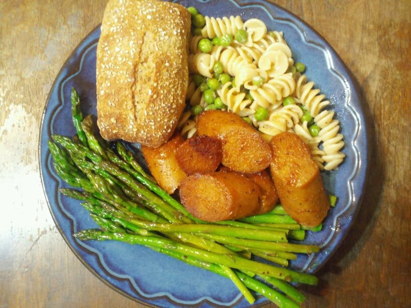 looking down at a blue plat that has pasta, a roll, asparagus and some kind of sauteed sausages on it