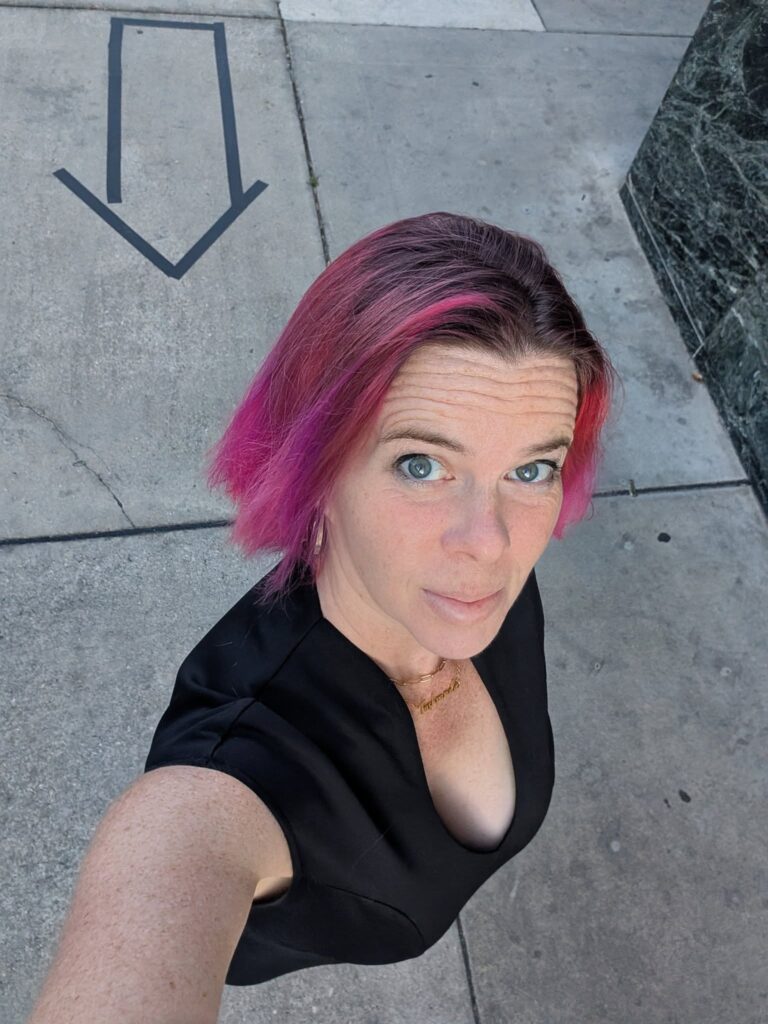 selfie looking down at a woman with pink hair and a black dress and behind her on the pavement is an arrow drawn in black lines pointing towards her back