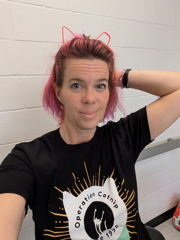 a person with pink hair and a cat ears headband is striking a pose in a black t-shirt that says Operation Catnip